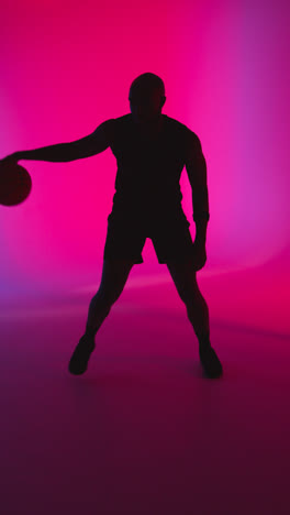 Vertikale-Videostudio-Silhouette-Eines-Männlichen-Basketballspielers,-Der-Vor-Rosa-Und-Blau-Beleuchtetem-Hintergrund-Dribbelt-Und-Den-Ball-Hüpft